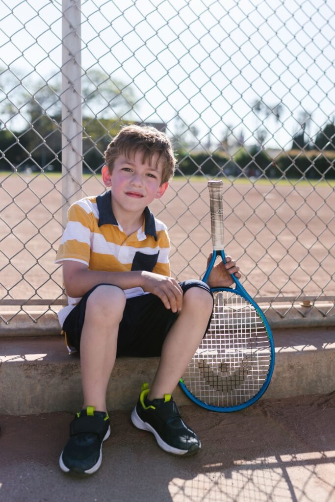 A smiling young boy in casual attire sitting outside with a tennis racket by a chain-link fence.